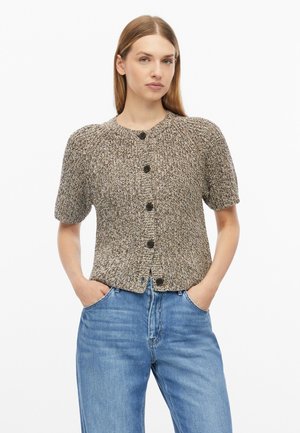 Jeune femme aux cheveux raides porte un cardigan en tricot à manches courtes boutonné et un jean bleu, debout les mains dans les poches.