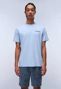 Light blue cotton t-shirt with short sleeves and round neck, featuring a small black logo on the left chest. Smooth texture, relaxed fit.