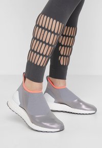 Šedé sportovní slip-on boty s hladkým povrchem, síťovaným svrškem a jasně oranžovými akcenty, spárované s černými legínami s výřezy.