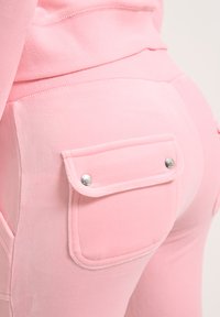 Un pantalon de survêtement rose au toucher doux, avec deux poches arrière ornées de boutons-pression argentés et des détails de surpiqûres contrastées.