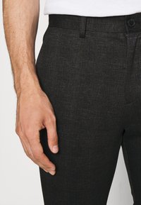 Pantalon à carreaux noirs avec une coupe sur mesure et un tissu texturé. Comprend une fermeture par boutons et une main visible posée sur le côté.