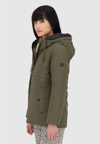 Parka verde oliva con cappuccio, chiusura con zip, tasche laterali, fodera interna arancione e dettagli neri. Presenta un logo sul petto e sulla manica.
