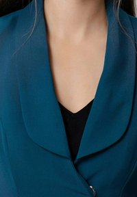 Gros plan sur une personne portant un blazer bleu-vert sur un haut noir à col en V, montrant le décolleté et une partie du col.