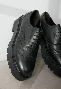 Chaussures brogues en cuir noir avec une finition texturée, présentant des détails perforés, des bords surélevés et des semelles épaisses en caoutchouc noir.