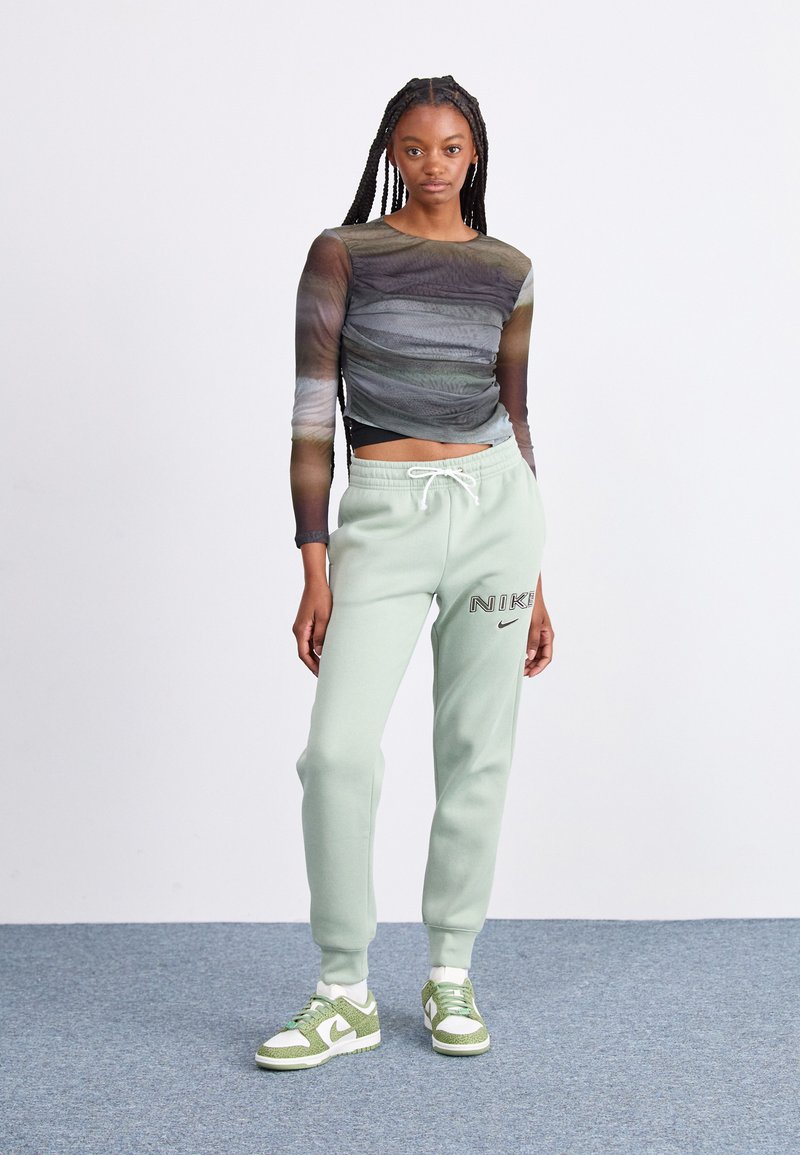 Haut à manches longues noir et vert avec texture froncée ; pantalons de sweat mint vert affichant le logo "Nike" ; baskets vertes et blanches avec des accents texturés.