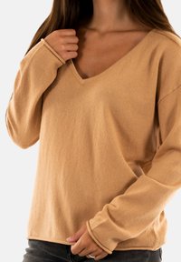 Maglione beige con scollo a V e maniche lunghe, realizzato in un tessuto morbido, con una vestibilità rilassata e polsini arrotolati, abbinato a jeans scuri.