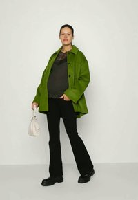 Casaco oversized verde com botões grandes, blusa preta, calças pretas flare e botas pretas robustas. Segura uma pequena mala branca.