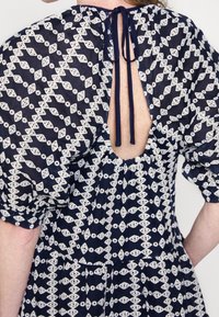 Femme porte une robe bleu marine avec un motif en dentelle géométrique blanche et un dos ouvert noué par un nœud au niveau du cou.