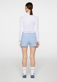 Vit långärmad tröja, ljusblå shorts, vita strumpor med logotyp; modell som bär handske och sneakers; minimalistisk design, figursydd silhuett.