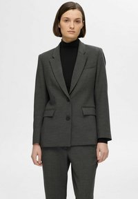 Blazer gris ajusté avec un col cranté, deux poches avant, fermeture à un bouton et texture lisse, porté sur un col roulé noir.