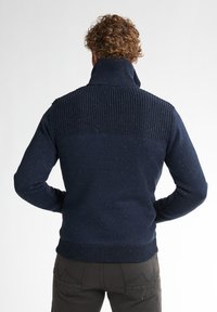 Marineblauwe gebreide trui met een hoge kraag. Gelaagd bovenstuk, gladde onderkant en kleurvlekken doorheen. Geribbelde manchetten en zoom.