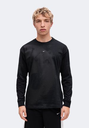 Млад мъж с къса, къдрава руса коса, носещ черна блуза с дълги ръкави Nike и черен панталон Nike, стои пред неутрален фон.