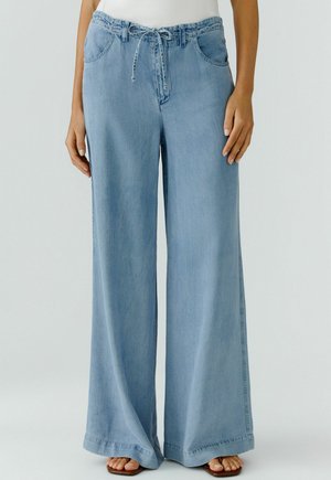 Femme portant un pantalon en denim bleu clair à jambes larges avec poches avant, taille à cordon de serrage, et sandales plates marron.
