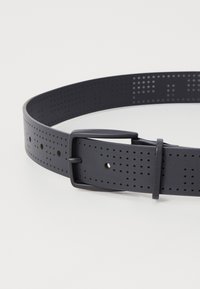 Μαύρης τρύπιας ζώνης από υλικό παρόμοιο με καουτσούκ, με ορθογώνια μεταλλική αγκράφα και πολλαπλές τρύπες ρύθμισης για ευελιξία μεγέθους.