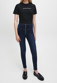 Černé bavlněné tričko s bílým nápisem "CALVIN KLEIN JEANS"; kombinováno s vysokým pasem tmavě modrých džínových kalhot s zipovým uzávěrem a kapsami.
