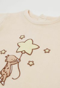 Un combinaisons en velours pêche avec un girafe brodée en brun tenant un ballon en forme d'étoile jaune, avec de petites étoiles jaunes comme accents.