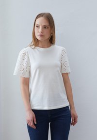 White cotton t-shirt with short puff sleeves featuring embroidered eyelet details and scalloped edges, paired with dark blue jeans.