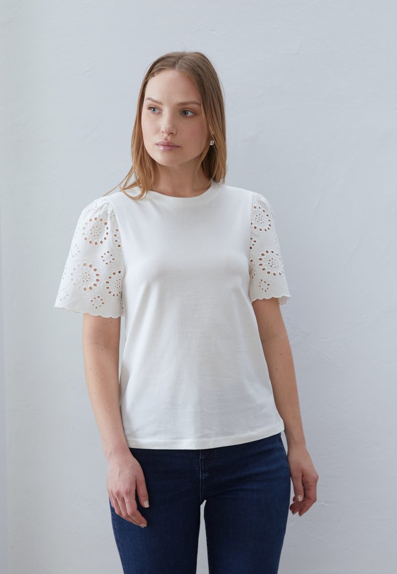 White cotton t-shirt with short puff sleeves featuring embroidered eyelet details and scalloped edges, paired with dark blue jeans.