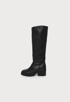 Botte en cuir noire montant jusqu’au genou, à bout arrondi et à gros talon bloc, sur un fond uni clair.