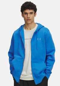 Sudadera azul con cremallera, capucha ajustable y bolsillos frontales, hecha de una tela suave. Lleva encima una camiseta blanca con texto impreso.