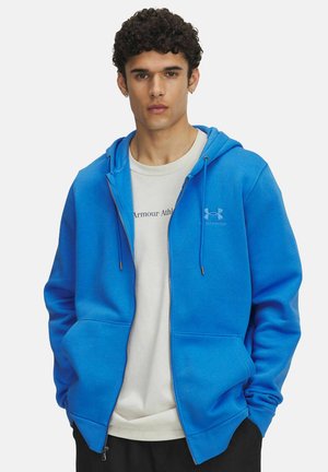 Blaue Zip-Hoodie mit Kordelzugkapuze und Fronttaschen, aus weichem Stoff. Über einem weißen T-Shirt mit bedrucktem Text getragen.