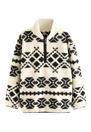 Person iført en sort og hvid mønstret fleece-pullover, burgunderfarvede leggings og sorte og pink løbesko, stående udendørs ved en stenmur.