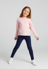 Młoda dziewczyna z długimi włosami, uśmiechnięta, ubrana w różową bluzkę z długim rękawem, granatowe legginsy i białe trampki, stojąca z nogami rozstawionymi na jednolitym tle.