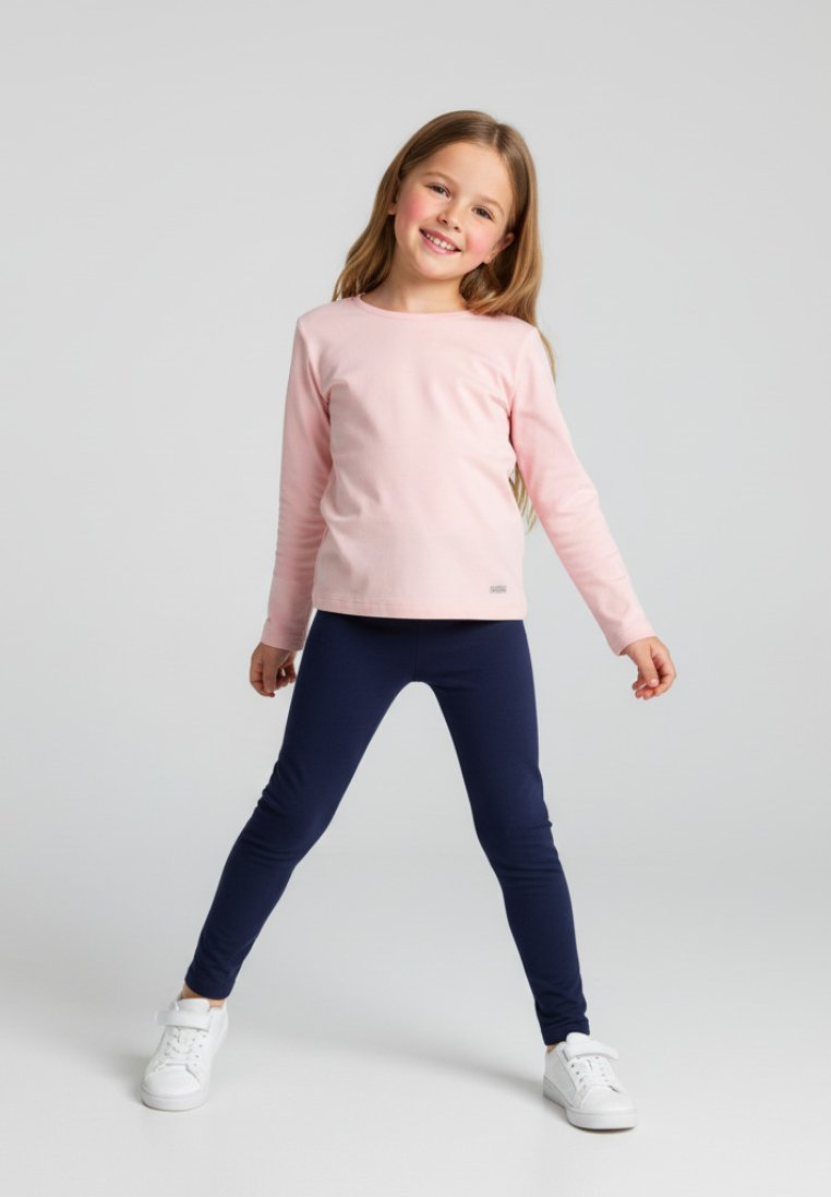 Młoda dziewczyna z długimi włosami, uśmiechnięta, ubrana w różową bluzkę z długim rękawem, granatowe legginsy i białe trampki, stojąca z nogami rozstawionymi na jednolitym tle.