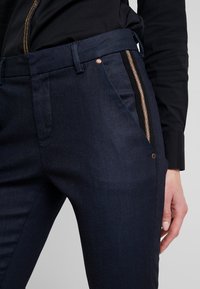 Pantalones azul marino con una textura suave, que cuentan con bolsillos con cremallera lateral y acentos verticales negros. Detalle de herrajes en cobre en la cintura.