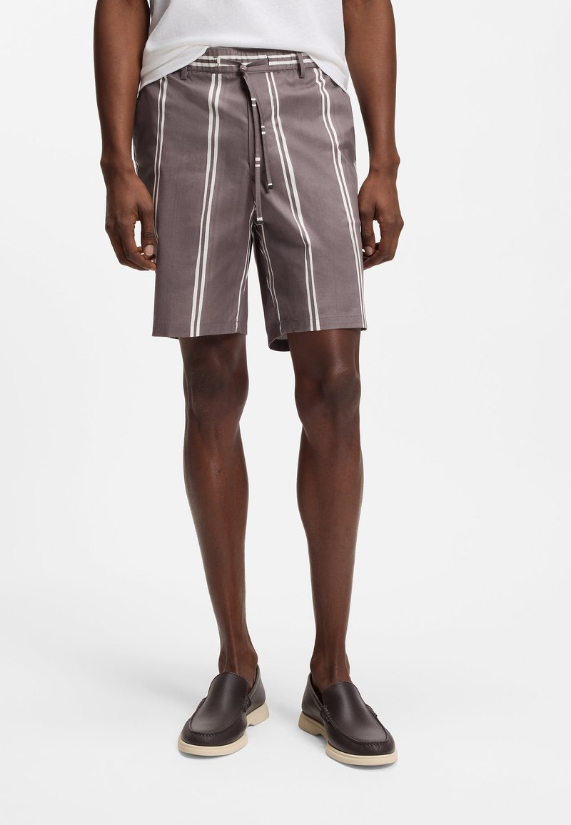 Uomo con pantaloncini grigi a righe verticali bianche, camicia bianca e scarpe slip-on marrone scuro su sfondo semplice.