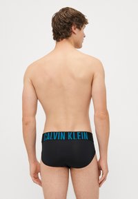 Fekete úszónadrág széles fekete derékkal, amelyen kék "CALVIN KLEIN" felirat található. Simább textúra, testhez álló forma és minimális fedettség jellemzi.