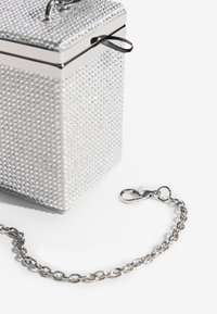 Ezüst színű téglalap alakú táska, melyet strasszkövek borítanak, kapcsos záródással, és egy fémes láncpánttal, kiemelve a textúrát és a csillogást.