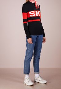 Schwarzer Pullover mit roten und weißen Streifen, versehen mit dem "SK"-Logo, kombiniert mit blauen Jeans und weißen Sneakern. Inklusive einem bedruckten Schal als Zubehör.