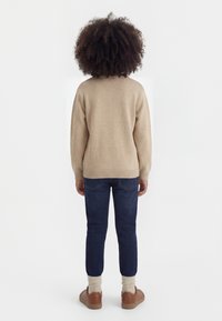 Bambino con capelli ricci che indossa un maglione beige, jeans scuri, scarpe color cuoio e calzini crema, in piedi di spalle su uno sfondo bianco.