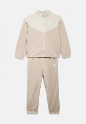 Téglabarna Nike melegítő egy cipzáros dzsekivel, amely világosabb bézs felsőrészű, és hozzáillő, szűkített nadrággal; mindkettőn elasztikus mandzsetták és logó található.