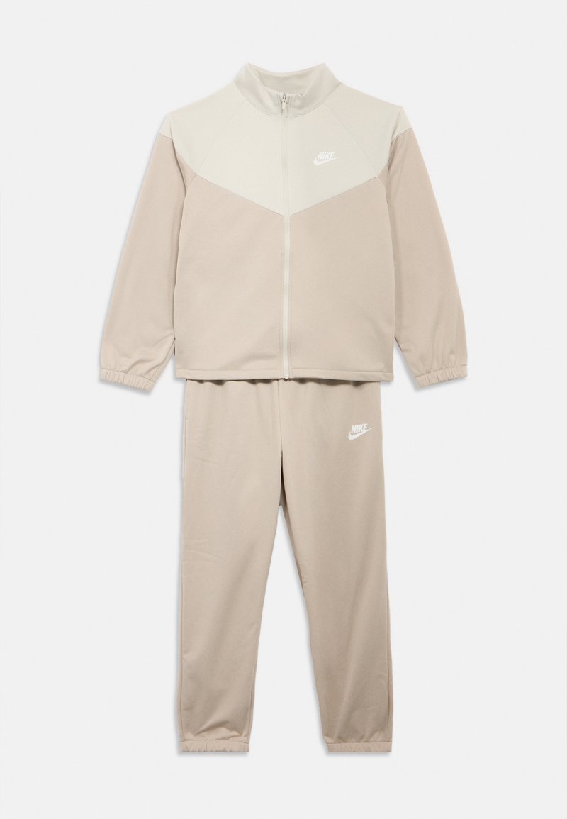Μπεζ φόρμα Nike με ζακέτα που κλείνει με φερμουάρ, με ανοιχτόχρωμη μπεζ επάνω περιοχή και ταιριαστό στενό παντελόνι· και τα δύο έχουν ελαστικές μανσέτες και λογότυπο.