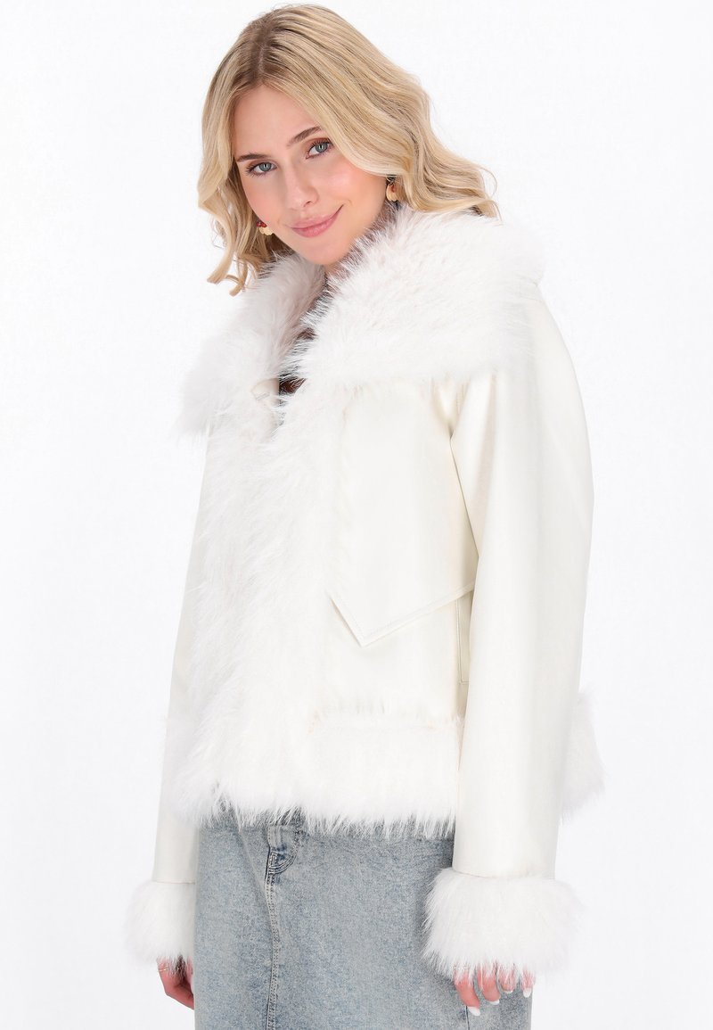 Giovane donna con capelli biondi che indossa un giubbotto bianco con bordi in pelliccia sintetica e jeans azzurri chiari, sorridendo dolcemente su uno sfondo bianco.