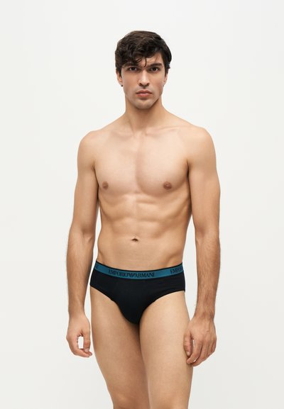 Jeune homme aux cheveux courts et foncés portant un slip noir à ceinture bleue, debout devant un fond clair uni.