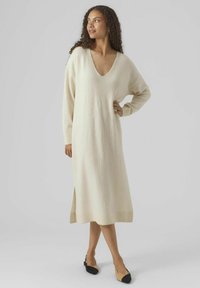 Robe beige en tricot à manches longues avec un col en V, longueur mi-mollet, fente sur le côté et poignets côtelés. Associée à des chaussures plates bicolores.