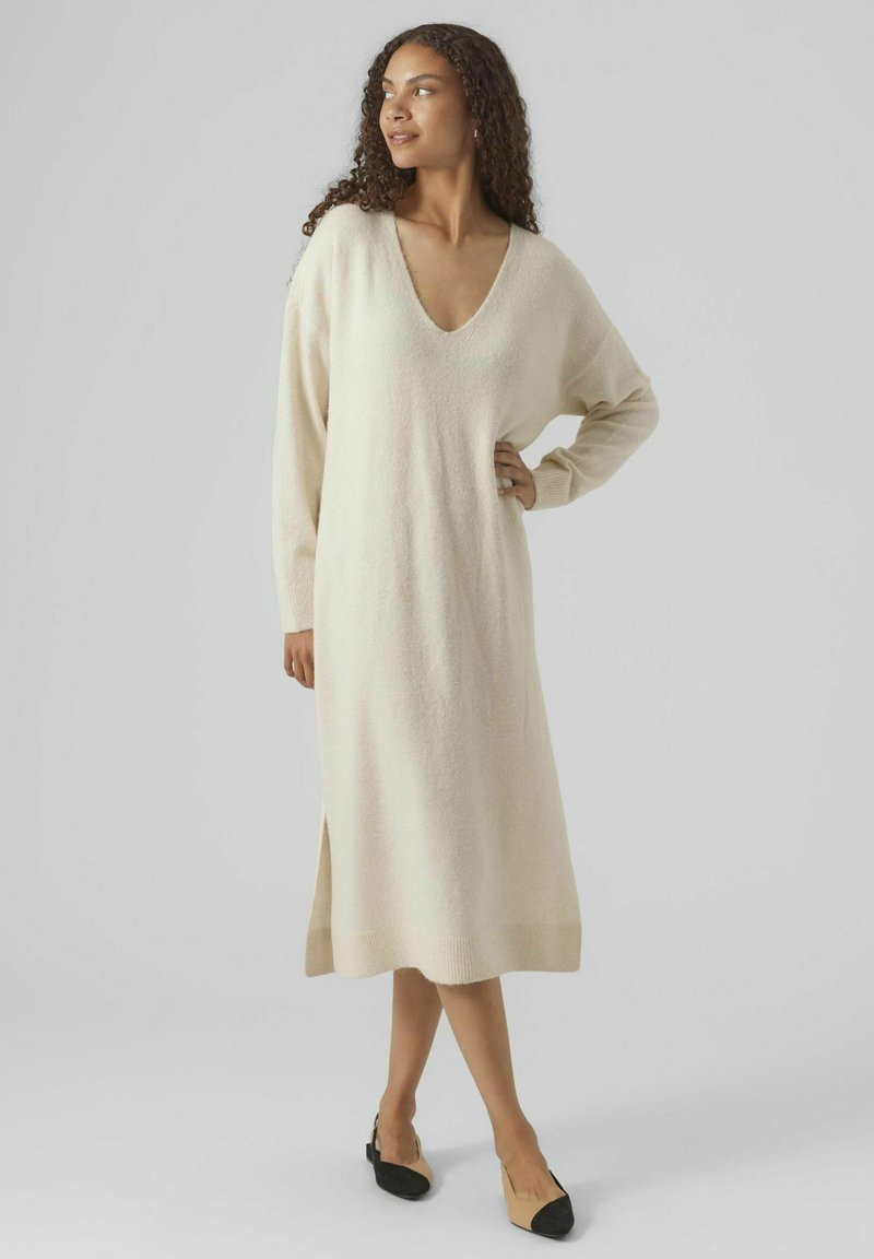 Robe beige en tricot à manches longues avec un col en V, longueur mi-mollet, fente sur le côté et poignets côtelés. Associée à des chaussures plates bicolores.
