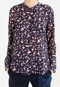 Långärmad rosa blus med mörk blommönster som bärs med mörka byxor som har en broderad blå drake på låret.