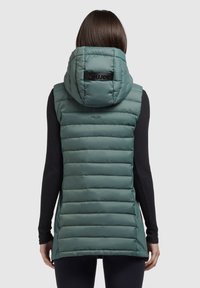 Kobieta w niebieskozielonej, pikowanej kamizelce bez rękawów z kapturem, ubrana w czarną bluzkę z długim rękawem, odwrócona tyłem, z prostymi brązowymi włosami widocznymi.