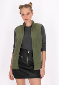 Gilet in pile verde oliva con zip frontale, abbinato a una maglia grigia aderente a maniche lunghe e una mini gonna nera in finta pelle con dettagli in zip.