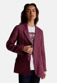 Femme portant un blazer violet sur une chemise blanche à motifs et un jean foncé, se tenant devant un fond clair et uni.