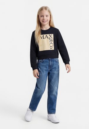 Ung pige med langt blond hår iført sort MAX&Co. sweatshirt, blå jeans og hvide sneakers, stående mod en hvid baggrund.