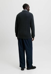 Pull en maille côtelée vert foncé à manches longues, associé à un pantalon bleu marine à rayures et des chaussures noires, vu de dos.