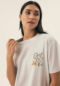 Biała bawełniana koszulka o luźnym kroju, z napisem "C'est la Vie" oraz pomarańczową grafiką w delikatnych odcieniach brązu i pomarańczy.
