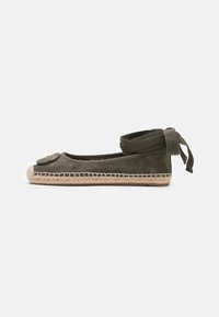 Chaussure plate en daim vert olive avec un bout arrondi, semelle en fibres naturelles tissées et liens en tissu à la cheville. Présente un emblème décoratif à l'avant.