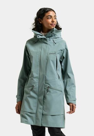 Femme portant un imperméable à capuche bleu clair avec des fermetures éclair à l'avant, debout et regardant légèrement vers la droite sur un fond uni.