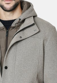 Chaqueta gris con patrón de espiga y capucha, que presenta un cierre frontal con cremallera y broches. Tela suave y texturizada con un interior forrado.